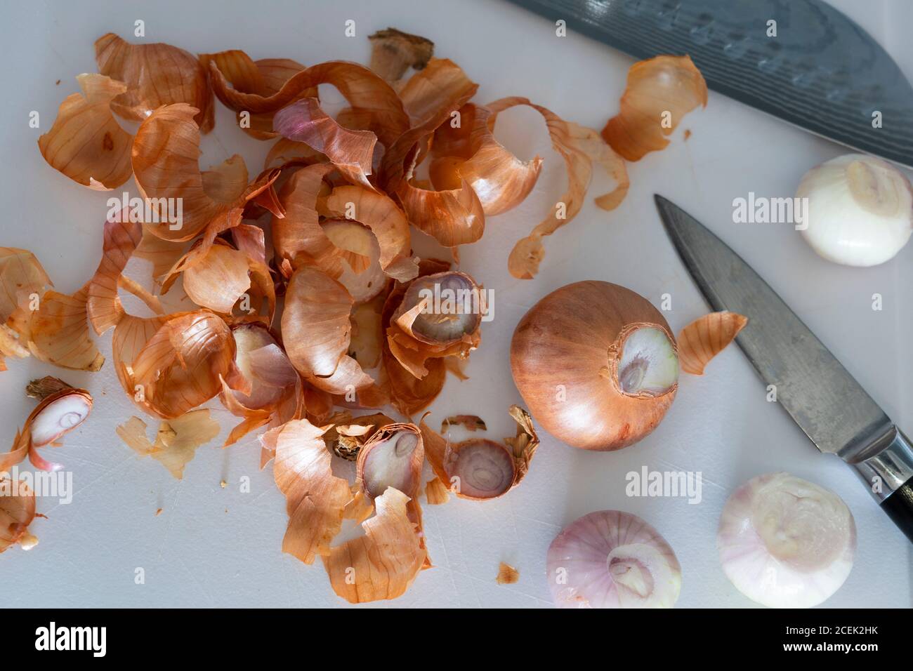 Peeled shallot onions with discarded onion skins on a white plastic chopping board with a Japanese knife Stock Photo