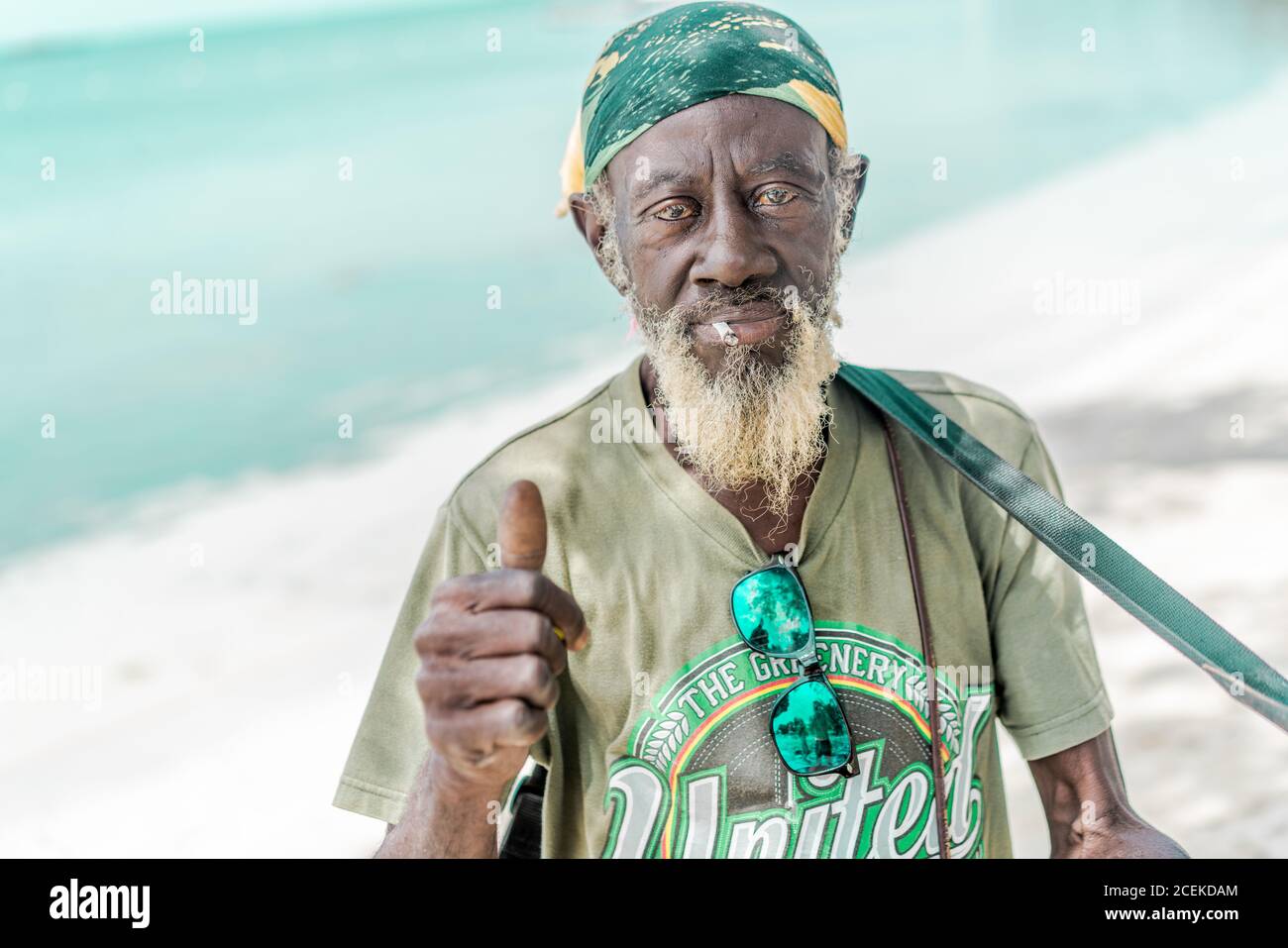 Senior Jamaican male with cigarette looking at camera and showing thumb-up gesture while standing near sea Stock Photo