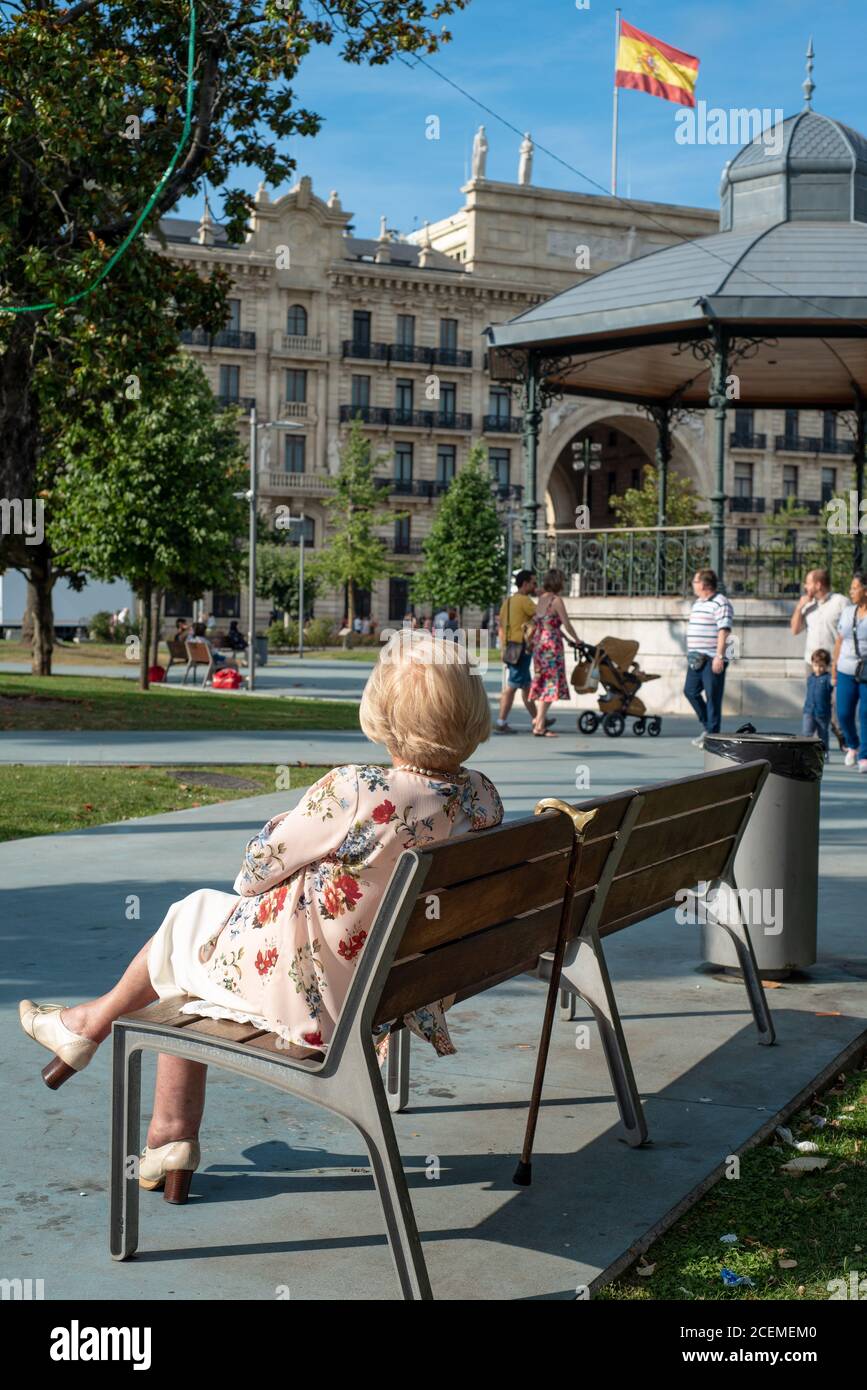 Back view of a middle-class blonde woman sitting on a wooden bench in Paseo Pereda. Sunny afternoon street scene in Santander, Cantabria. July 2019. Stock Photo