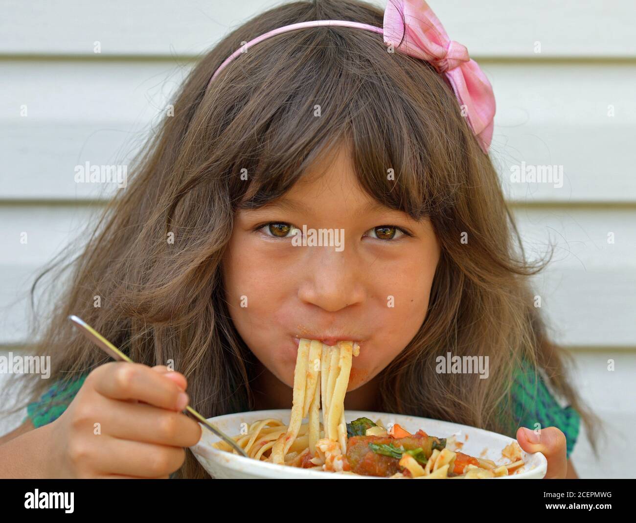 Hungry mixed-race cute little girl (East Asian / Caucasian) with pink bow knot in her hair eats dry noodles with a fork from a white ceramic bowl. Stock Photo