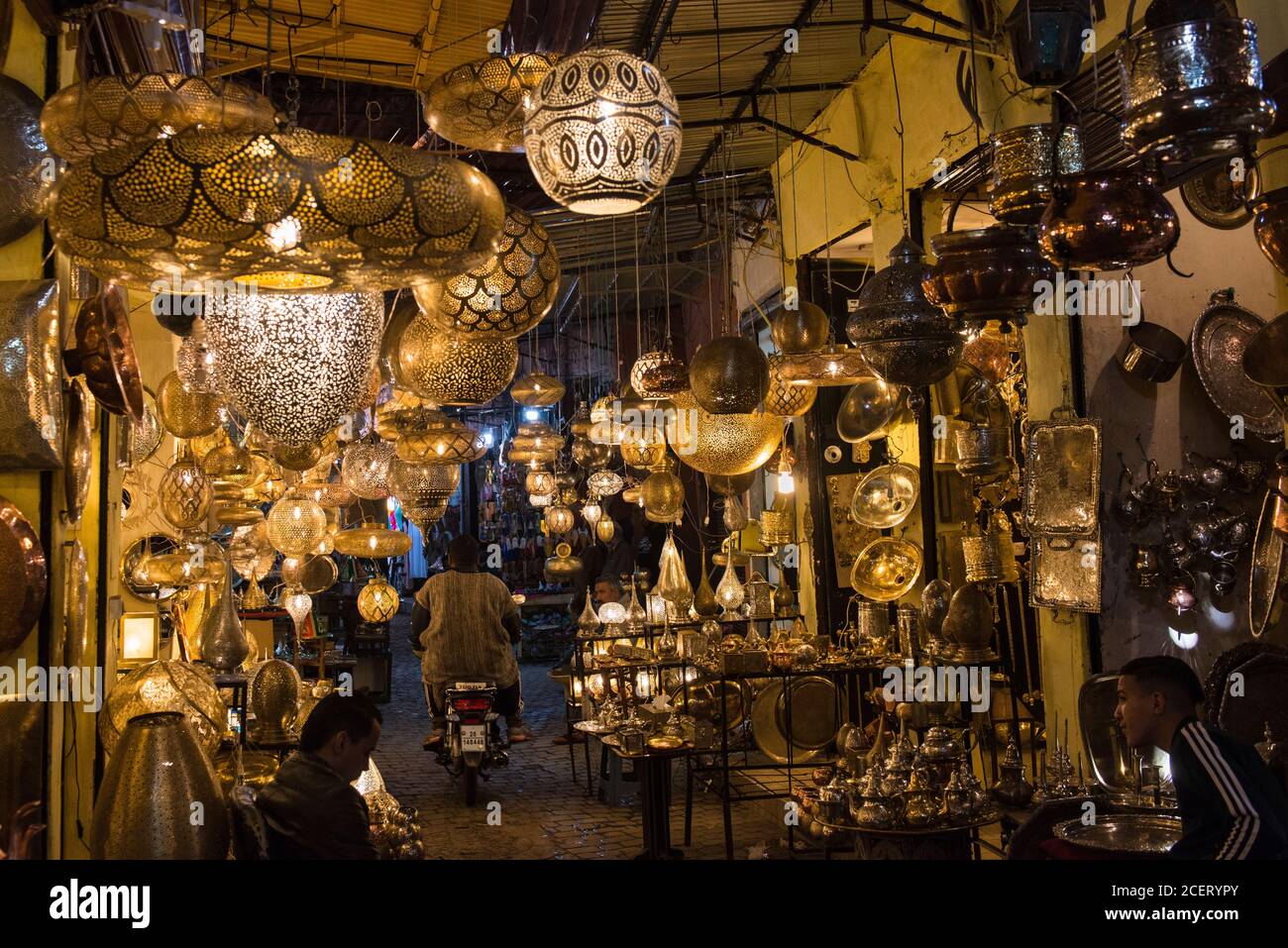 A motorbike rider passes between stalls selling Moroccan lampshades and ceiling lights  in the souk inside the Medina, Marrakesh Stock Photo