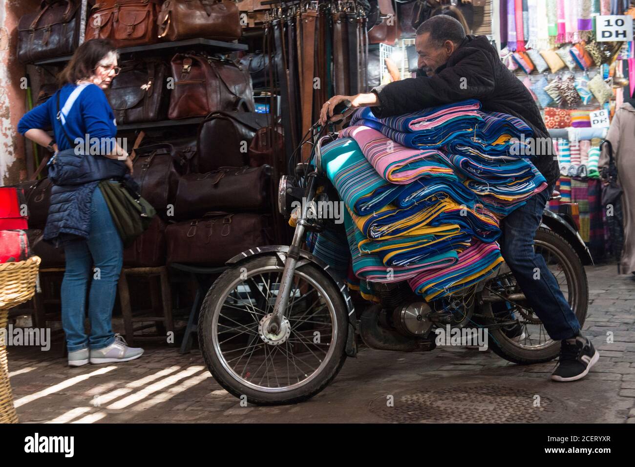 Don''t tell health and safety! Tourist watches a man riding his motorbike, overloaded with rolls of fabric, through the souk in Marrakesh Stock Photo