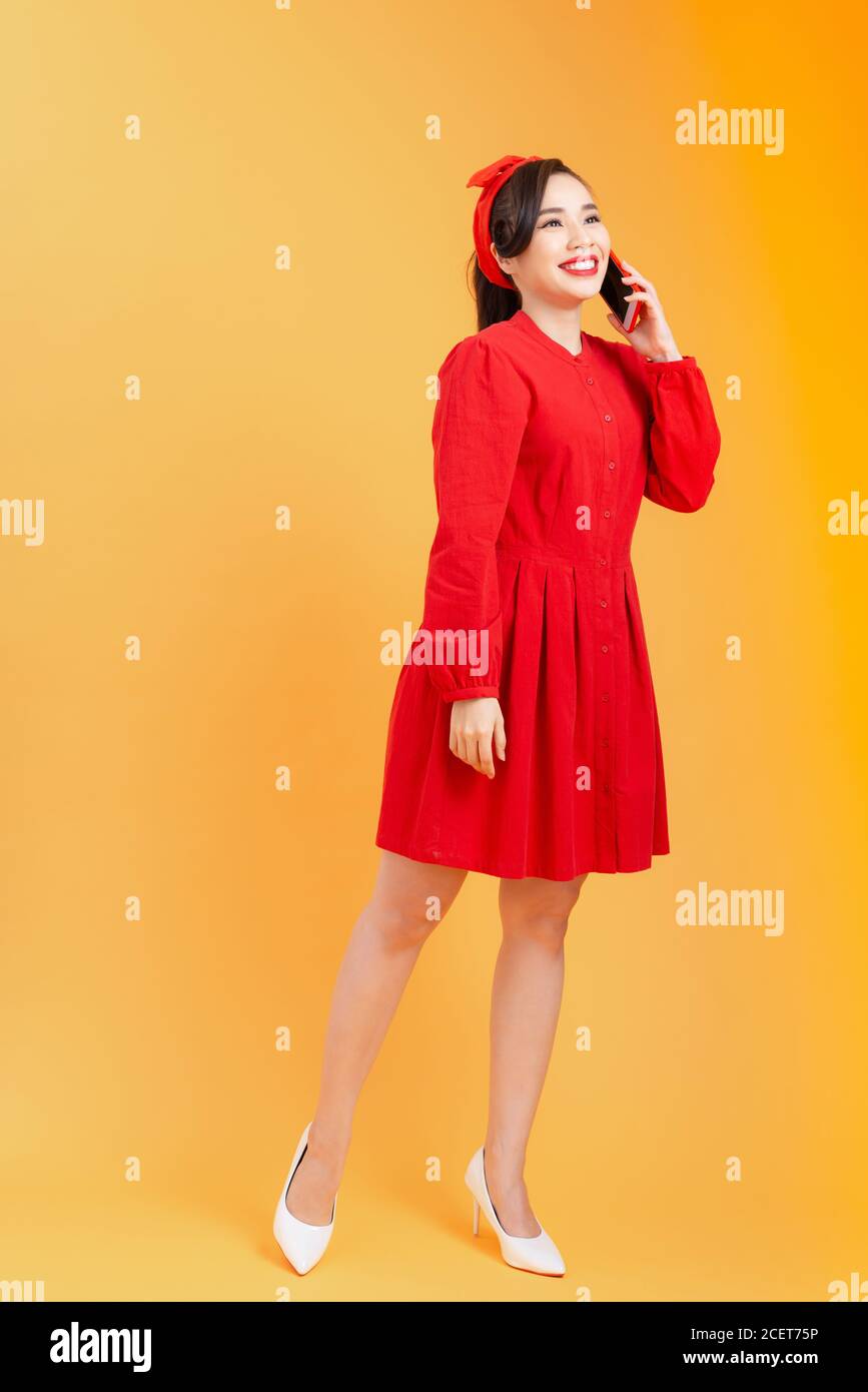 Cheerful young Asian woman in red dress talking on phone, standing over ...