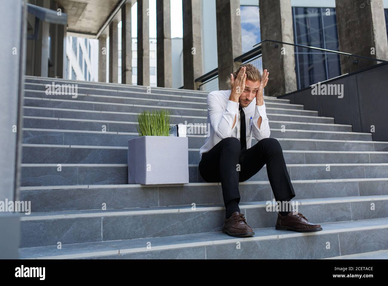 An unhappy young manager in a white shirt is fired from his job. A sad worker sits on the steps of a business center after layoffs. Crisis and Stock Photo