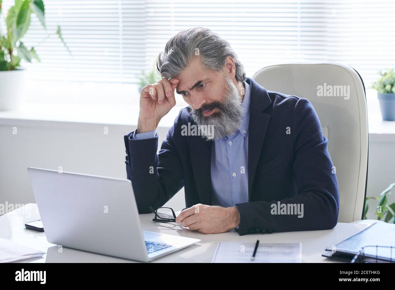 Portrait of concentrated mature male office worker wearing eyeglasses sitting at office desk and reading information on laptop Stock Photo