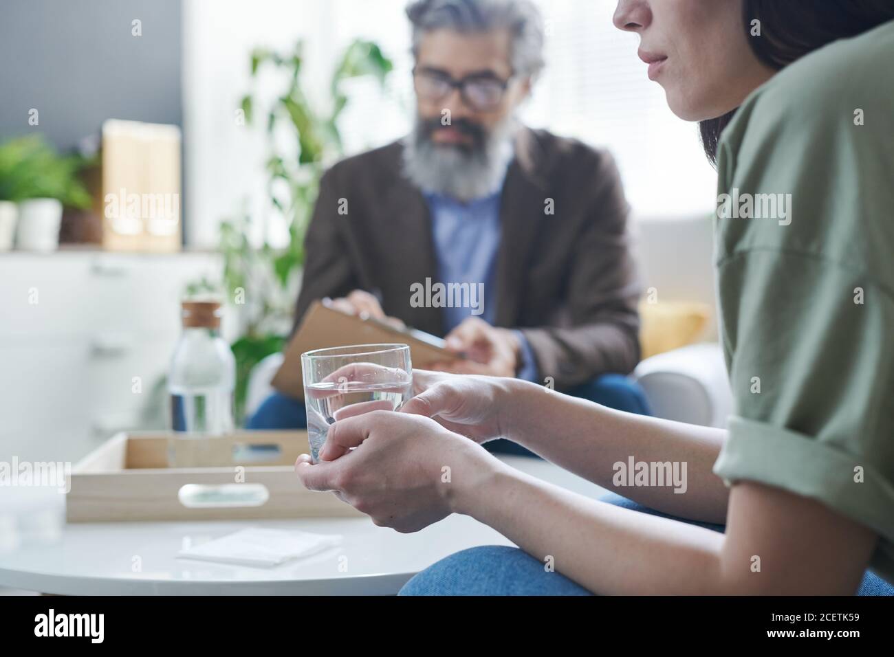 Low-spirited woman holding glass of water trying to calm down during therapy session at psychologist office Stock Photo