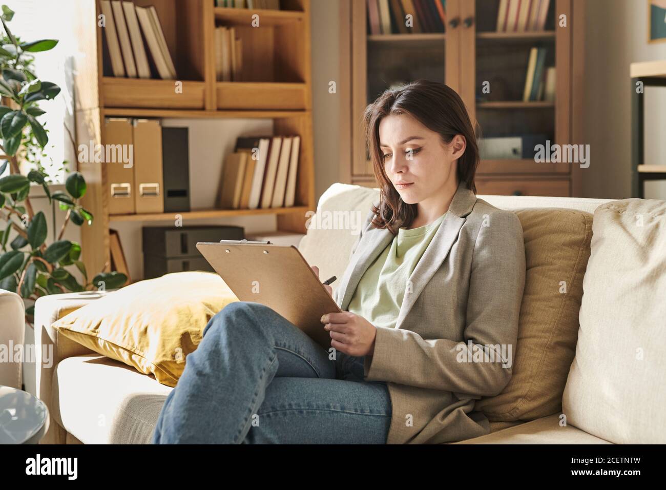 Attractive young woman sitting on sofa in modern office room holding clipboard making notes Stock Photo