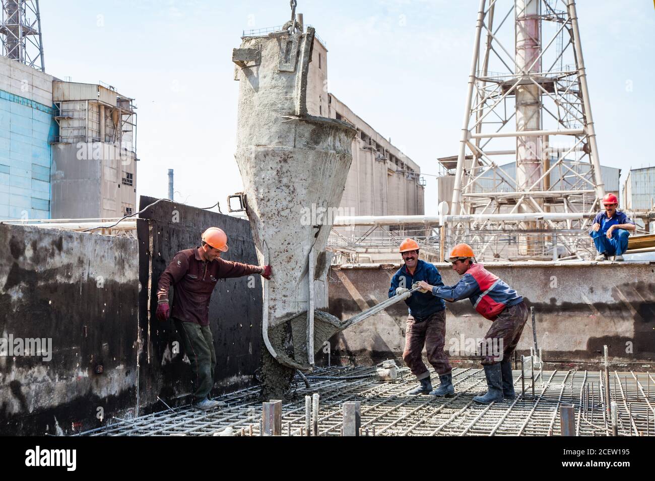 Old phosphate fertilizer plant in modernization. Workers with cement bucket filling of reinforcement of new building basement. On industrial building, Stock Photo