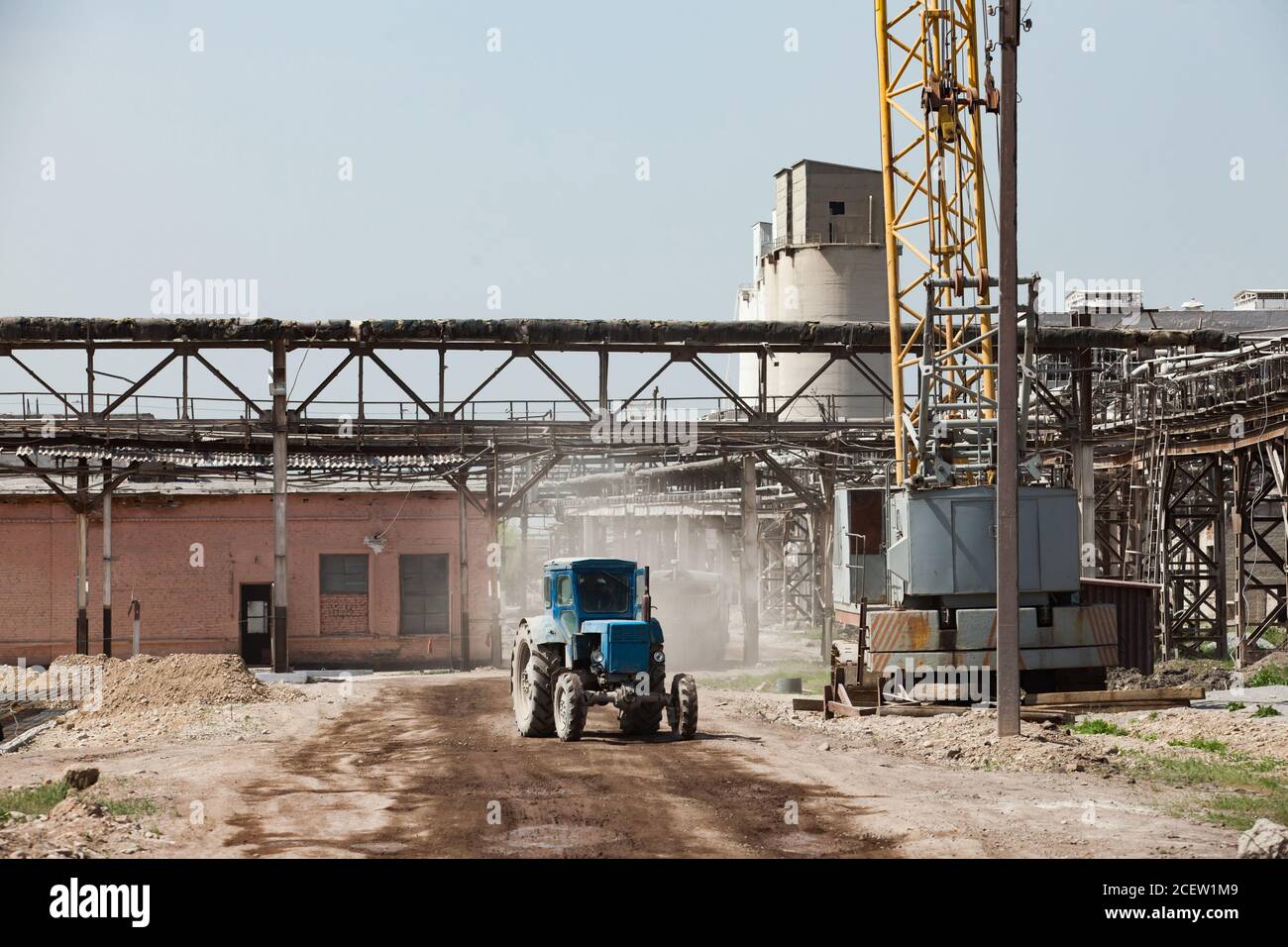 Grey pipelines and concrete construction and girder crane on grey sky. Tractor Belarus on the dusted soil. Old plant renovation. Stock Photo
