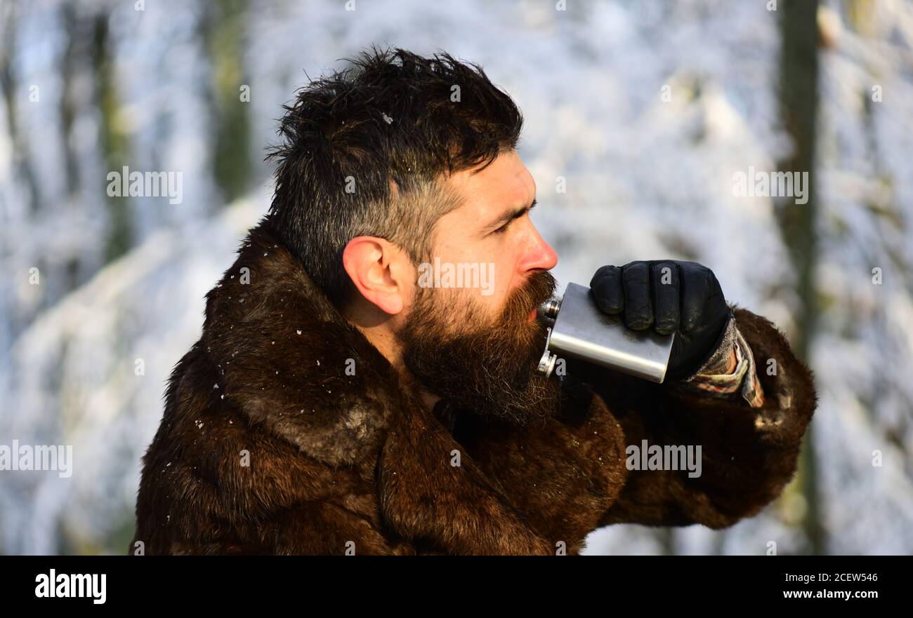 Macho with beard and mustache in forest. Guy on snowy nature park background, defocused. Brutal gamekeeper concept. Man in fur coat drinks from metal flask. Stock Photo