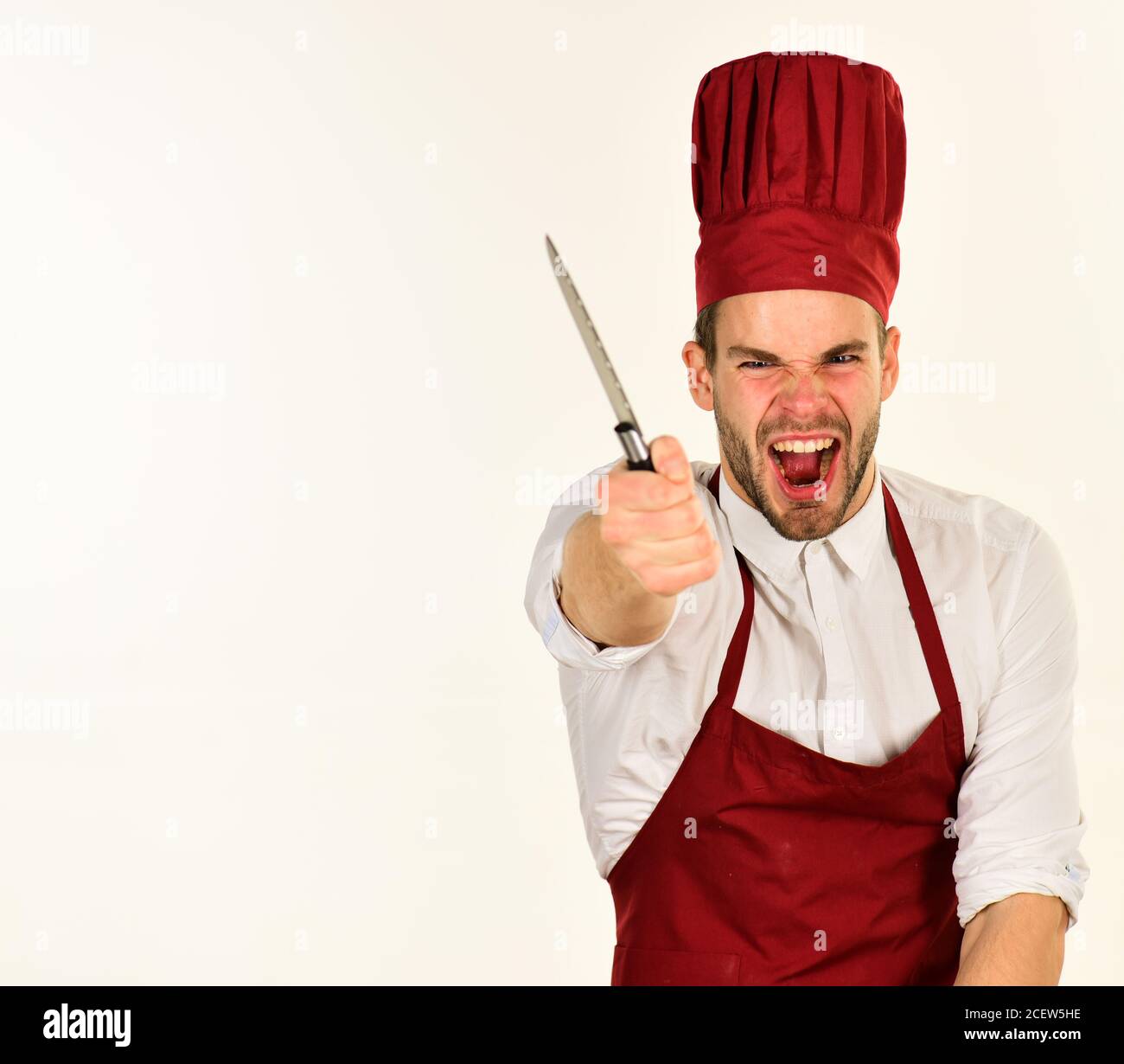 Cook works in kitchen. Man in cook hat and apron shows sharp blade forward in rage. Chef with angry face holds knife on white background. Food preparation and kitchenware concept. Stock Photo