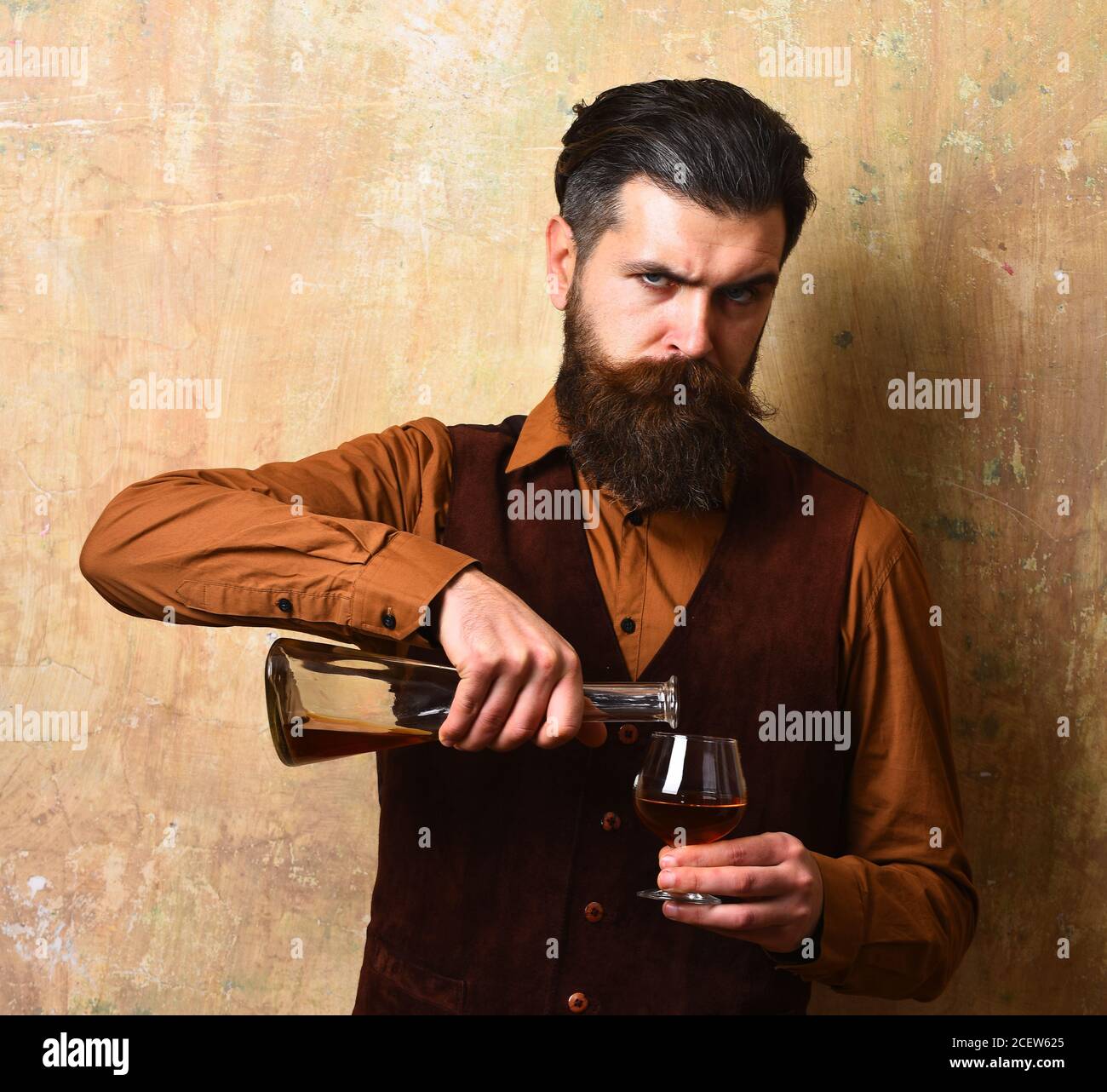 Guy pours cognac into glass from bottle. Man with beard and mustache holds alcoholic beverage on beige wall background. Service and catering concept. Macho with strict face drinks brandy or whiskey. Stock Photo