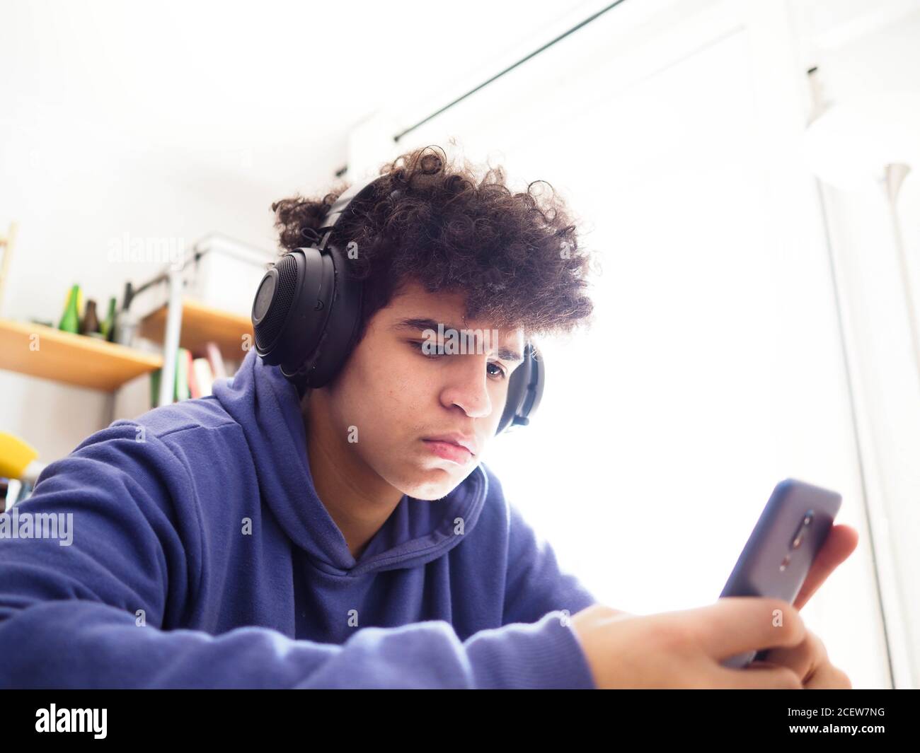 Curly young male in a purple hoodie and headphones using his phone while studying Stock Photo