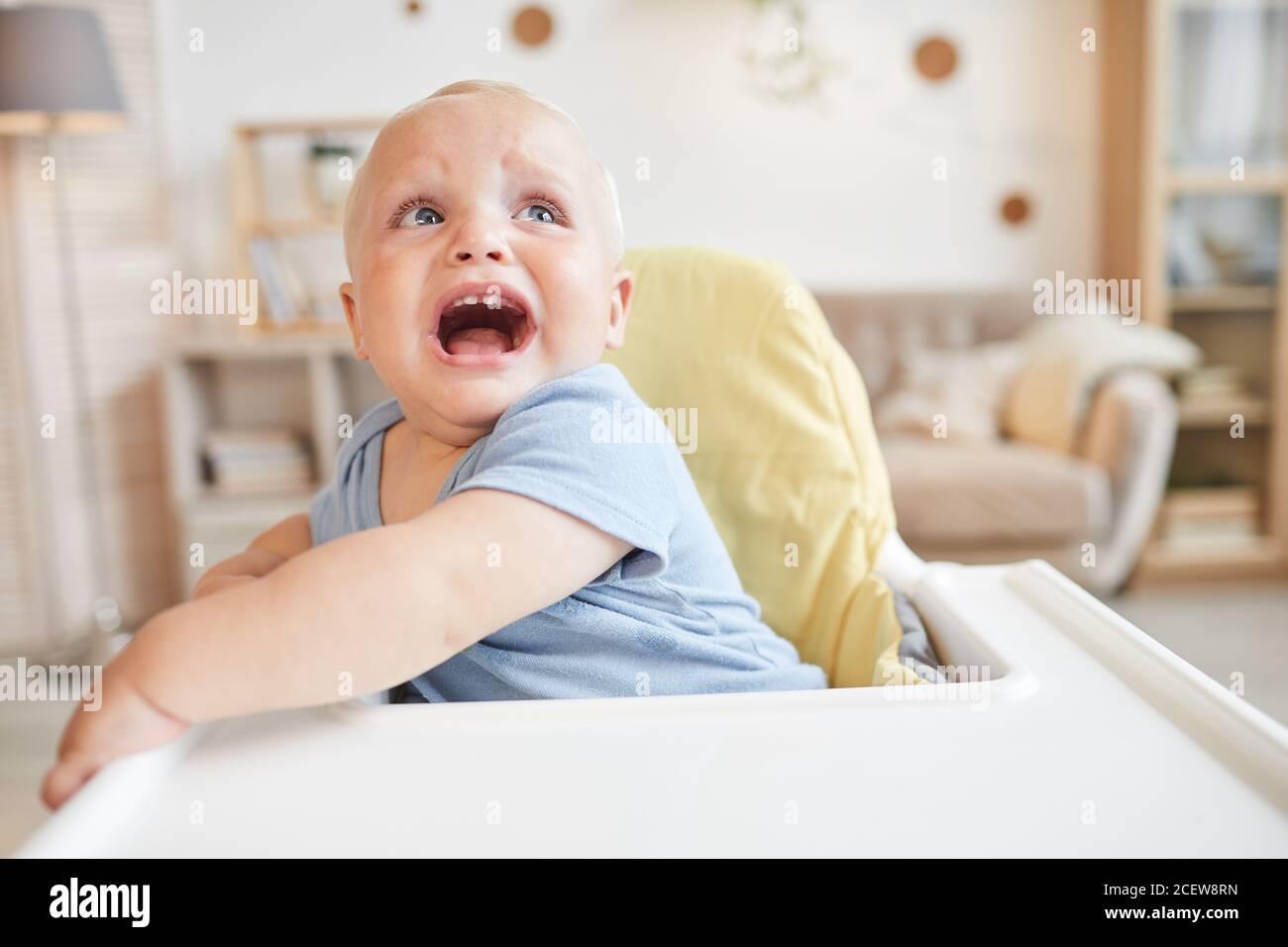 Horizontal medium close-up portrait of naughty baby boy sitting on high chair crying for something Stock Photo