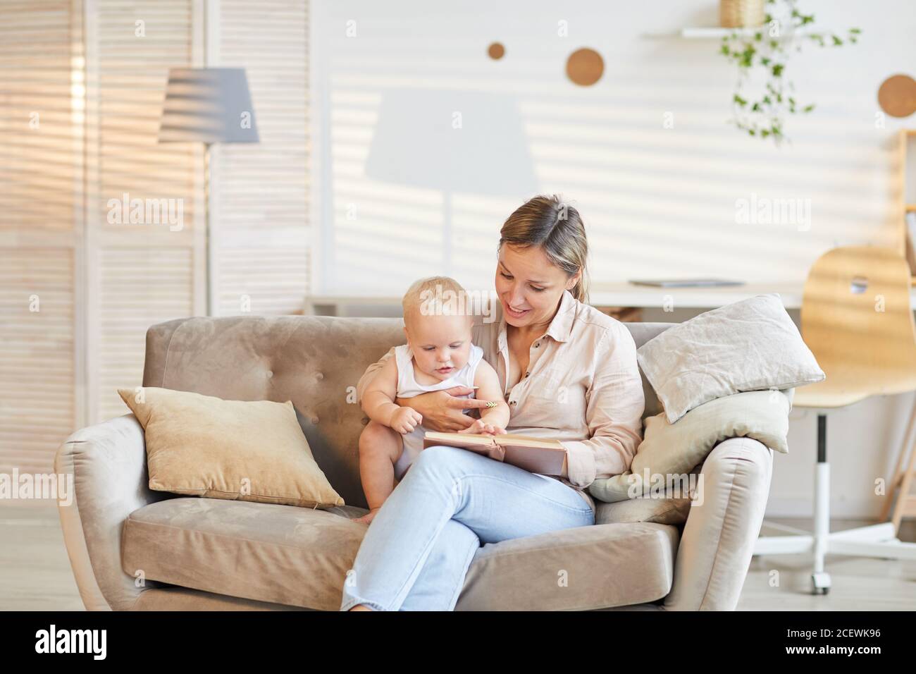 Modern young mother sitting on sofa in living room with her curious kid reading book, copy space Stock Photo