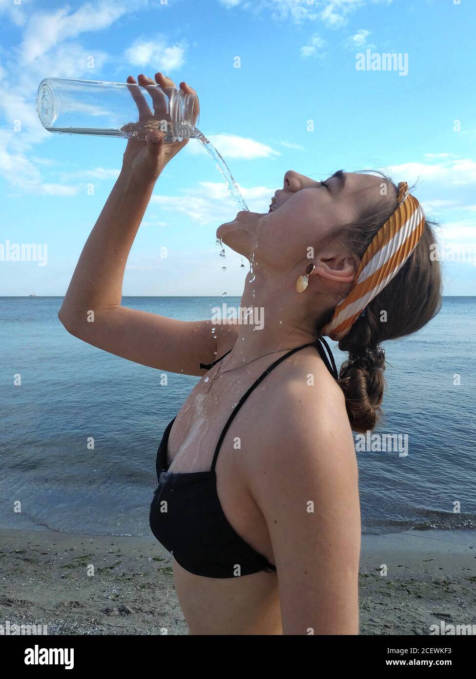Beautiful young woman in swimsuit drinks clean water from bottle, against blue teal sea and sky in sunny summer day on beach. Health care, healthy act Stock Photo