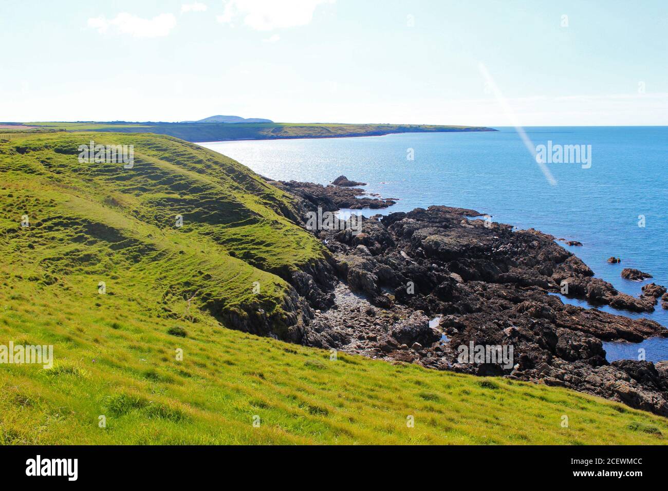 Beautiful scenery of the welsh coast, inc a green hill and rocky shore next to blue sea on a sunny day in Porth Colmen, North Wales Stock Photo