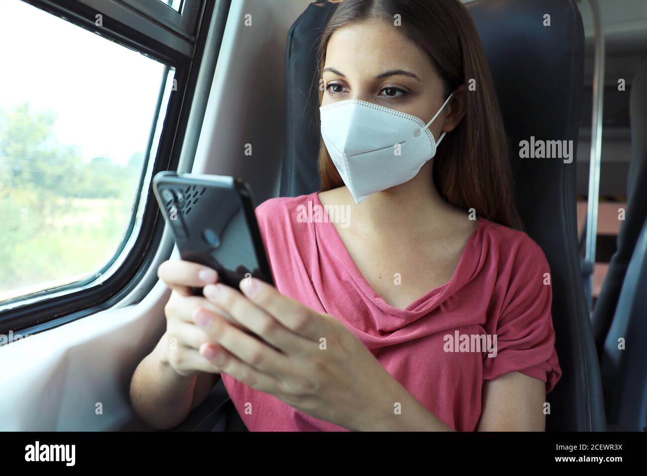 Relaxed woman with KN95 FFP2 face mask using smart phone app. Train passenger with protective mask traveling sitting in business class texting on mobi Stock Photo