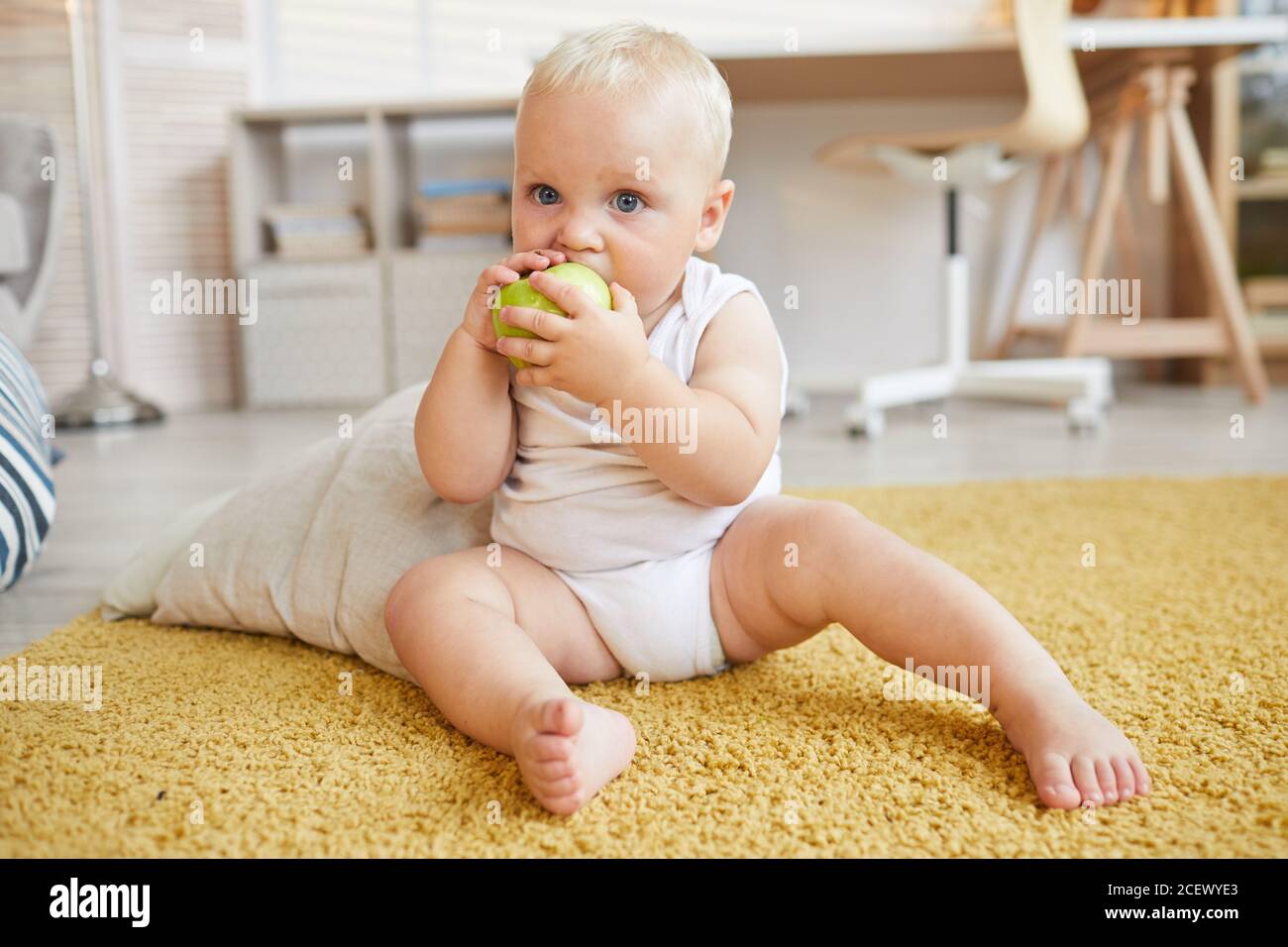 Horizontal full body shot of baby sitting on carpet biting and eating green apple, copy space Stock Photo
