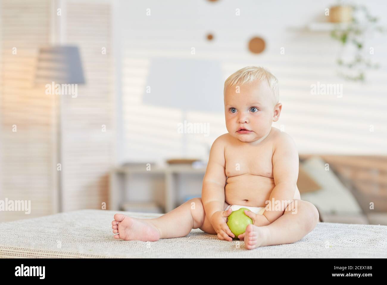 Cute little baby sitting on table in living room holding fresh green apple looking away, copy space Stock Photo