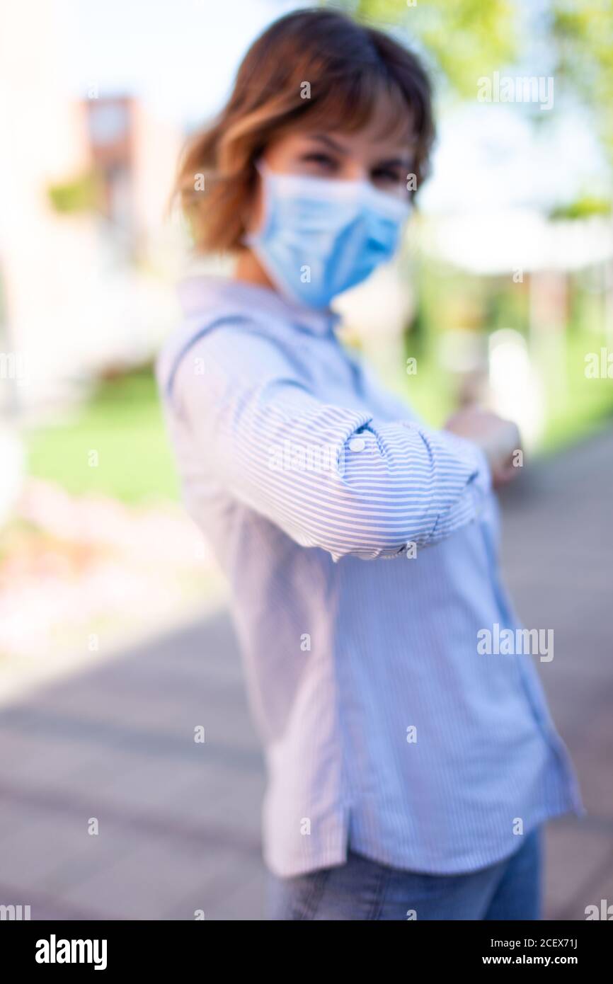 Young woman in mask greeting with elbow outdoors in park during pandemic, depth of field Stock Photo