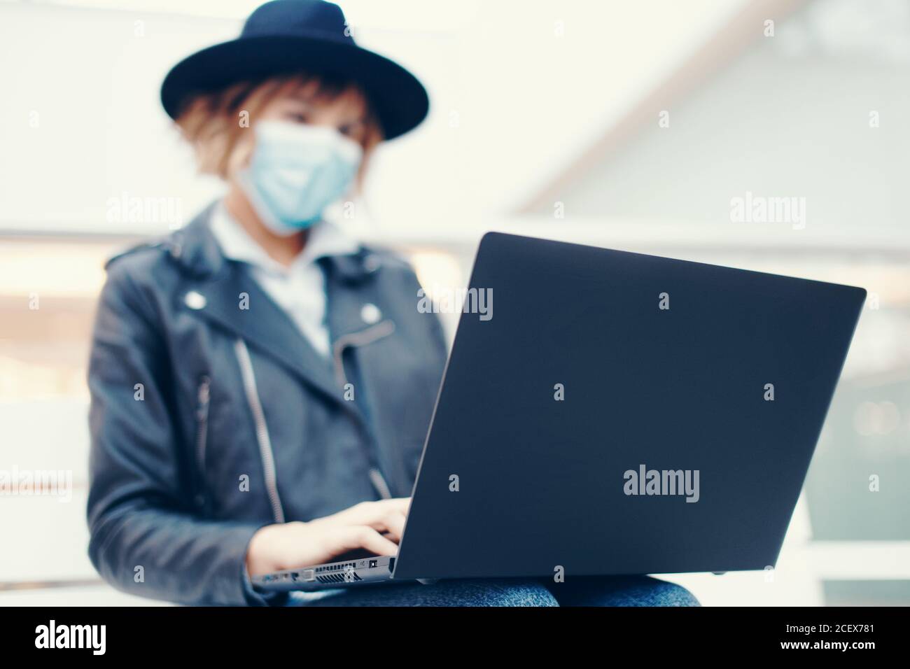 Young trendy woman in mask using laptop indoors, depth of field Stock Photo