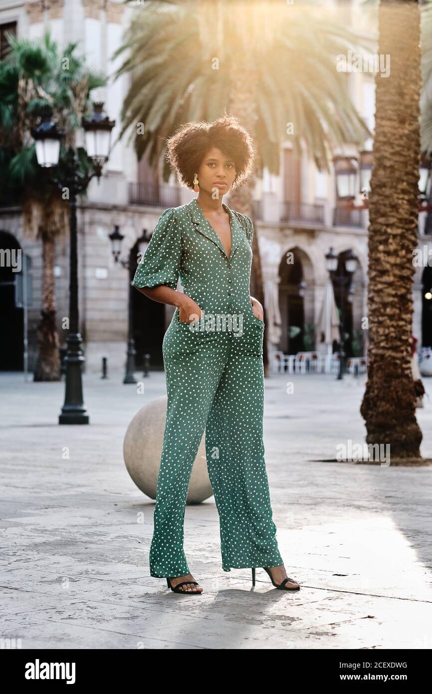 Young African American female with curly hair and stylish earrings wearing trendy green polka dot jumpsuit looking at camera while standing against blurred urban background Stock Photo