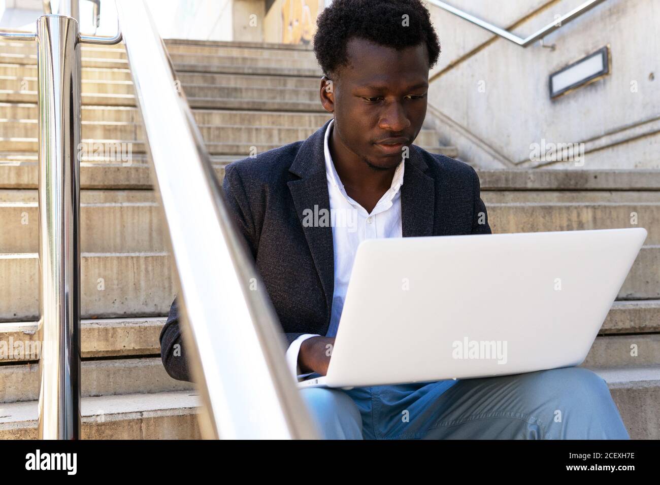 Serious African American male entrepreneur sitting on stone stairs in city and working on remote project while using modern netbook Stock Photo