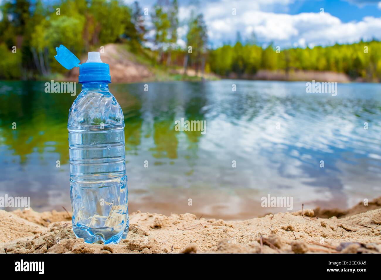 https://c8.alamy.com/comp/2CEXKA5/a-blue-plastic-bottle-with-an-open-lid-stands-on-a-sandy-beach-against-the-backdrop-of-a-wild-lake-background-2CEXKA5.jpg