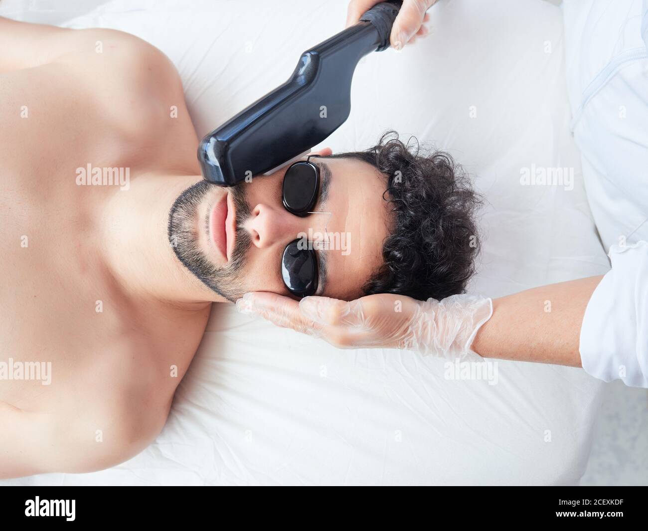 From above cropped faceless master using laser apparatus on male face removing beard face during procedure in salon Stock Photo