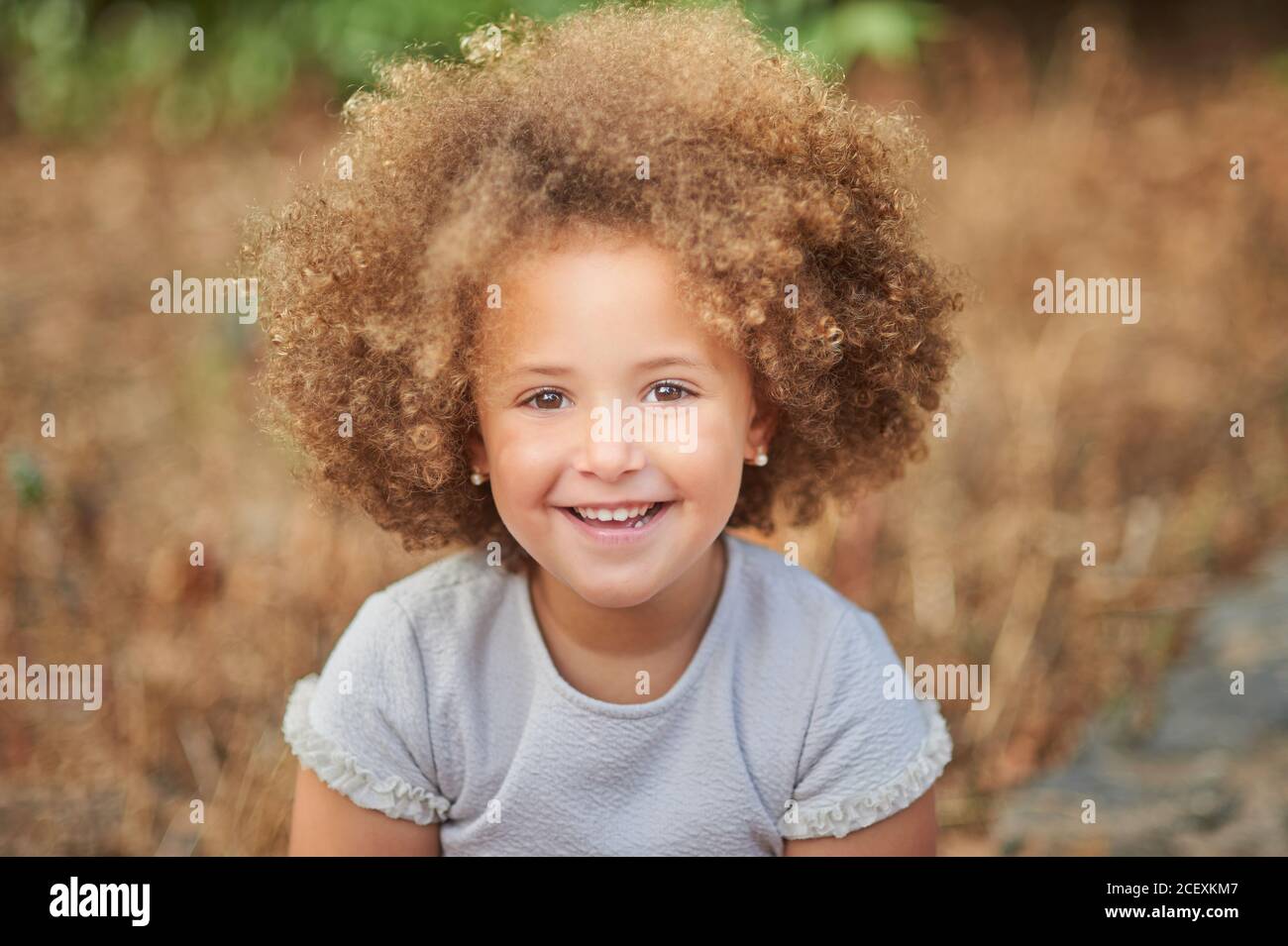 Adorable little girl with blonde Afro hair smiling and looking at camera while spending summer day in nature Stock Photo