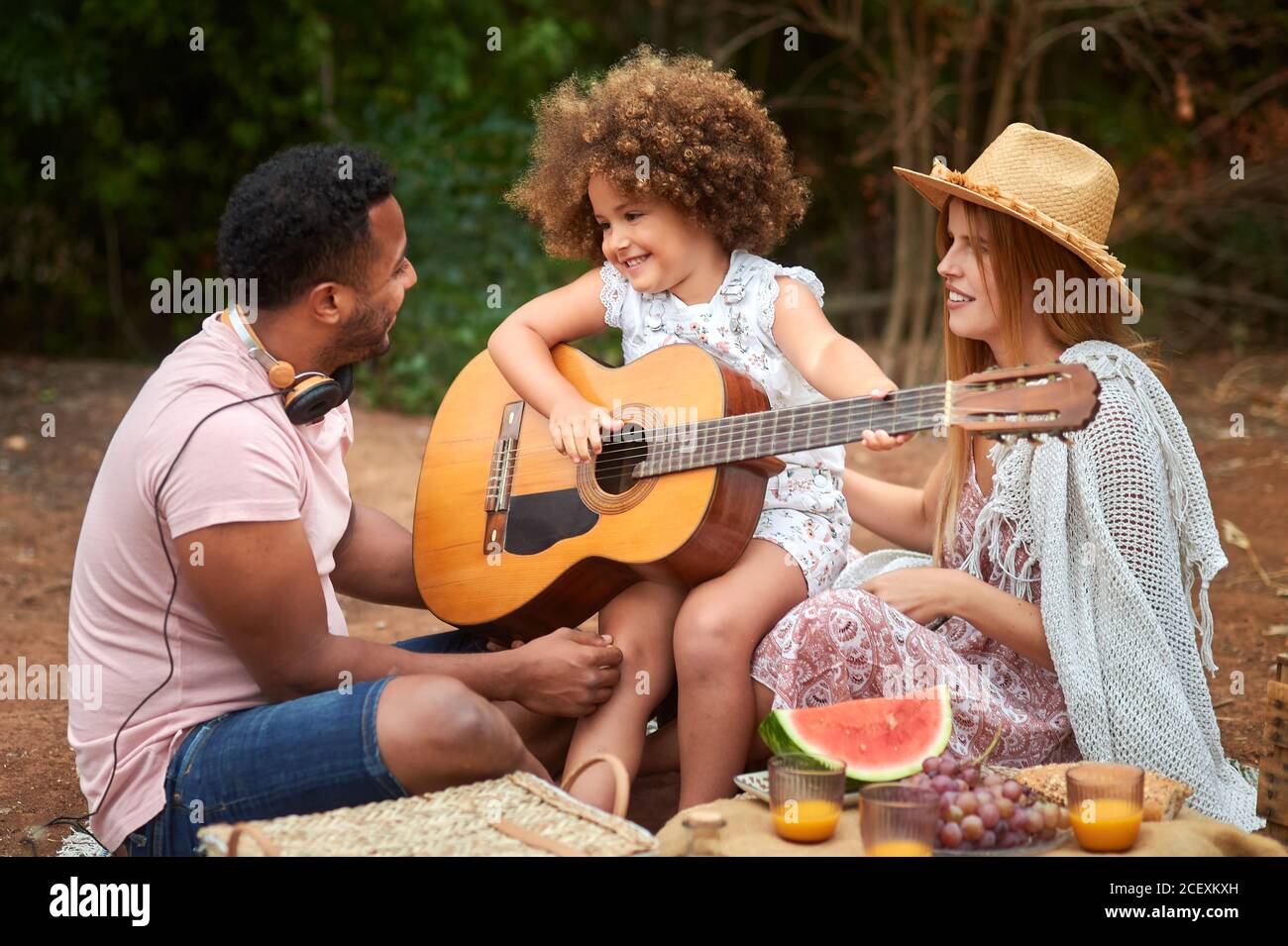 Cute curly haired little girl having fun and playing guitar during picnic with multiracial parents in summer day in nature Stock Photo