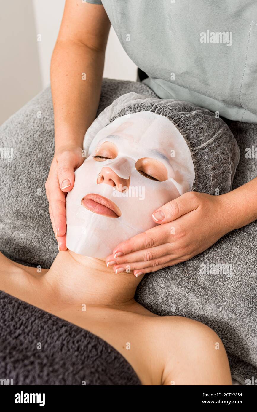 Crop anonymous cosmetician applying facial cloth mask on female client during rejuvenate skincare procedure in modern beauty salon Stock Photo