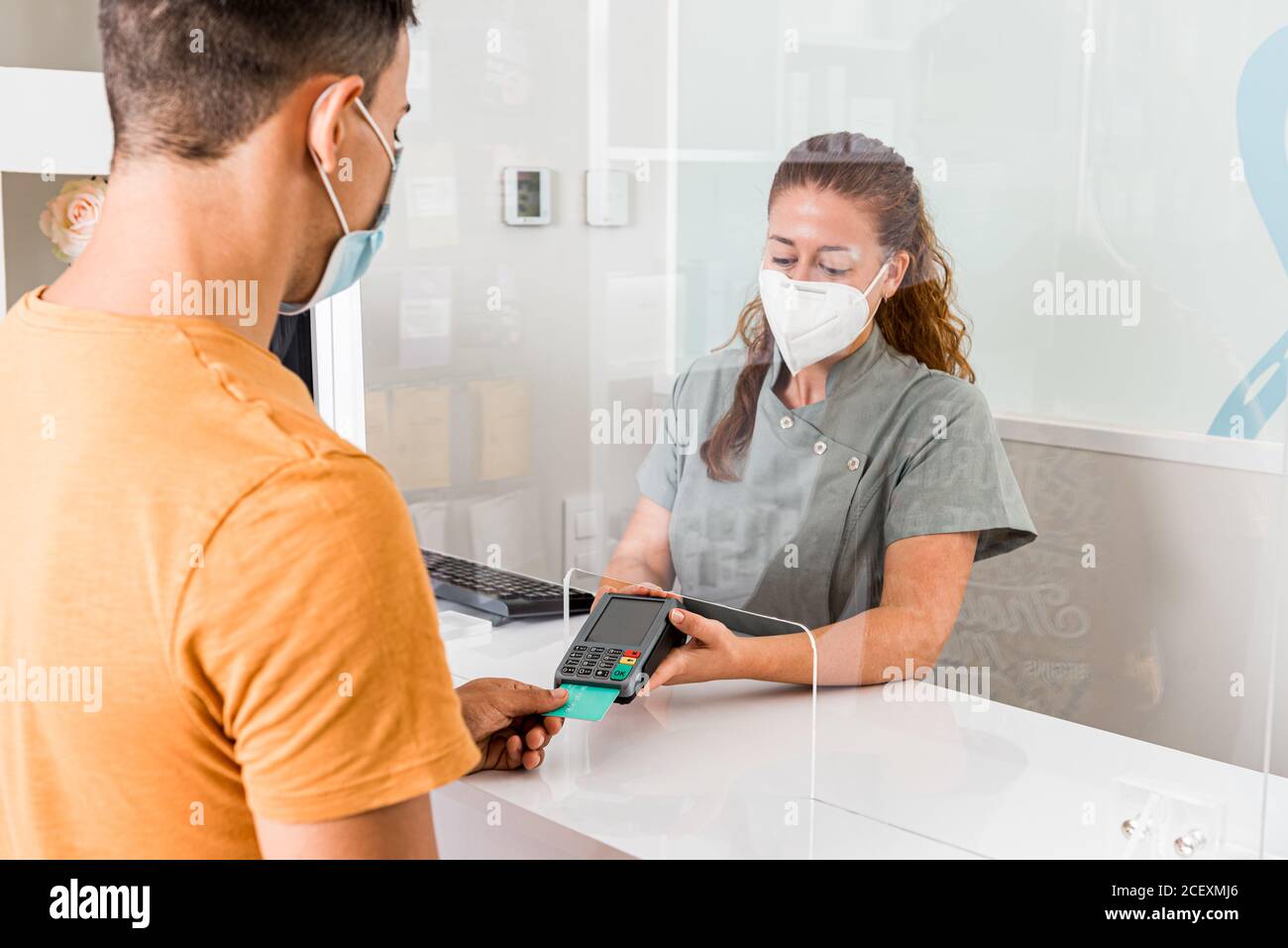 Young female specialist in protective mask holding card reader while male patient paying for treatment with credit card in modern clinic Stock Photo