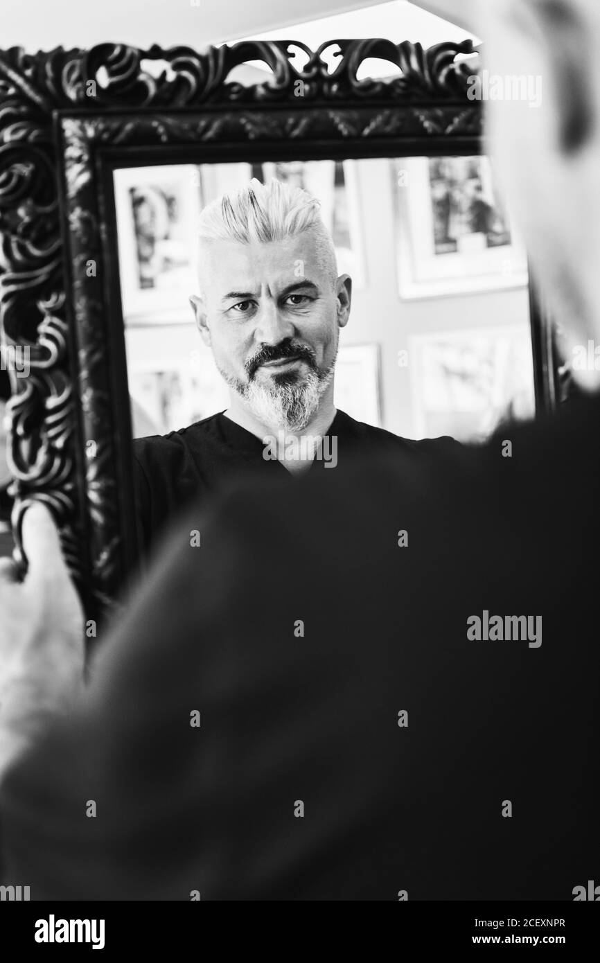 Confident brutal middle aged bearded male with gray hair in black clothes looking at reflection in framed mirror Stock Photo