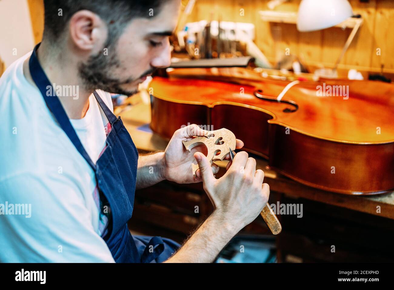 Side view of skilled young male artisan with knife carving wooden violin bridge while creating string instrument in workshop Stock Photo