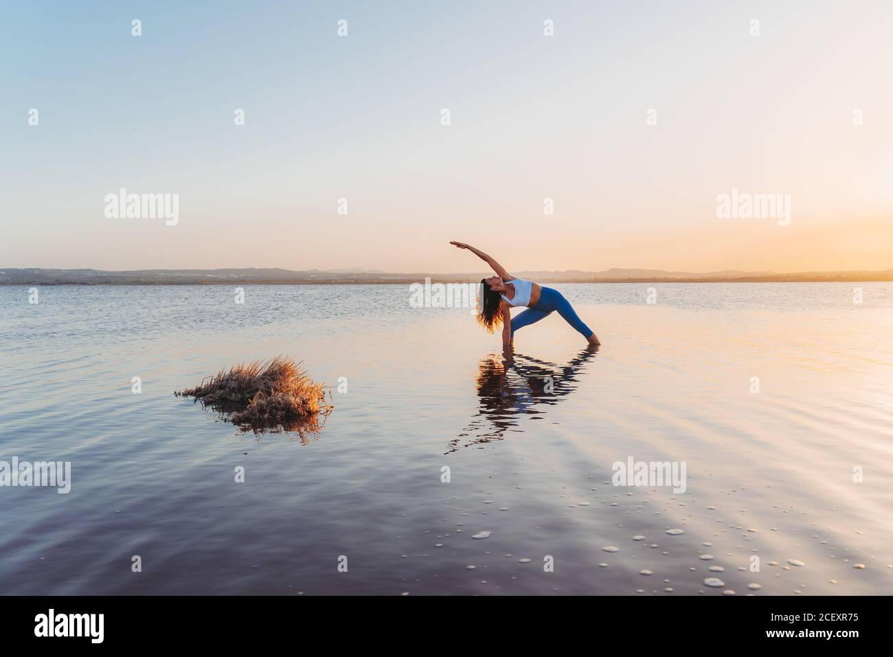 Full length of young slim female in activewear standing in extended side angle pose yoga in calm lake water during summer sunset Stock Photo