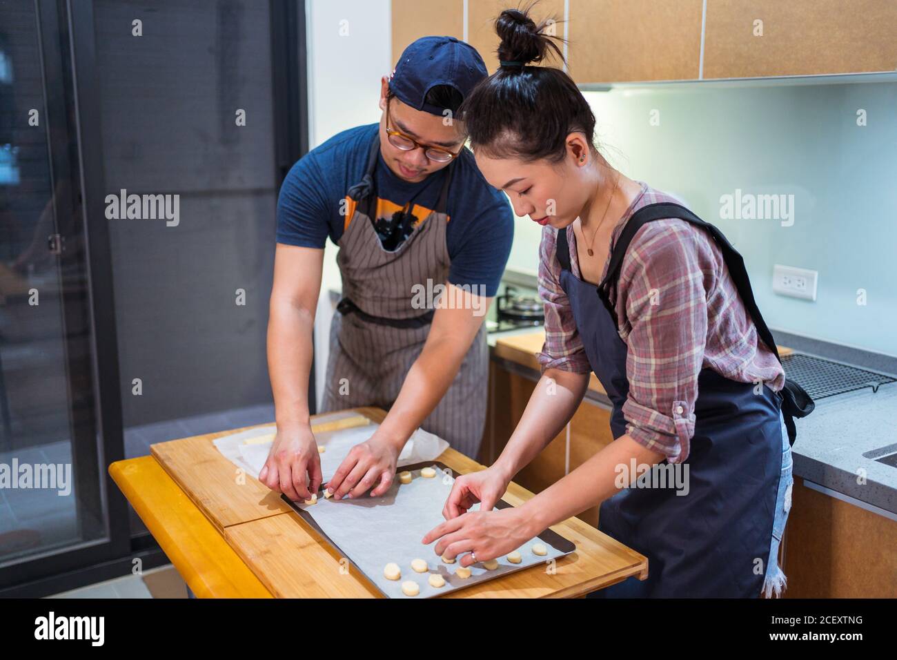 Young Asian man and Woman in aprons working at home bakery and placing cookies on baking pan with parchment Stock Photo