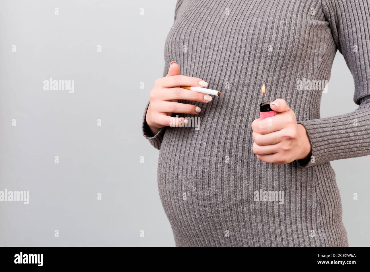 Close up of a cigarette and a lighter in pregnant woman's hands at gray background. Unhealthy