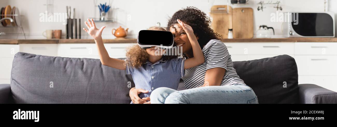 panoramic shot of young african american babysitter hugging girl in vr headset gesturing in kitchen Stock Photo