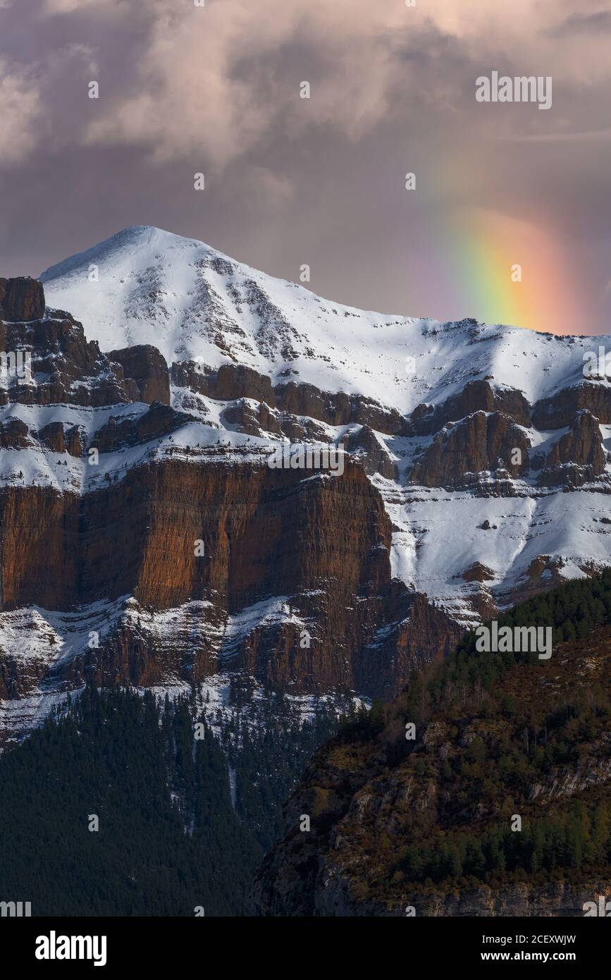 Spectacular landscape of mountain with snowy peak on slope under cloudy sky with rainbow Stock Photo