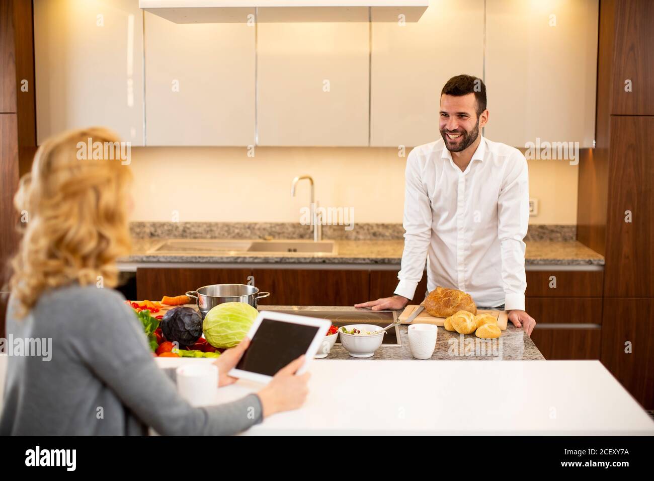 Young woman using tablet at the kitchen table while man preparing food Stock Photo