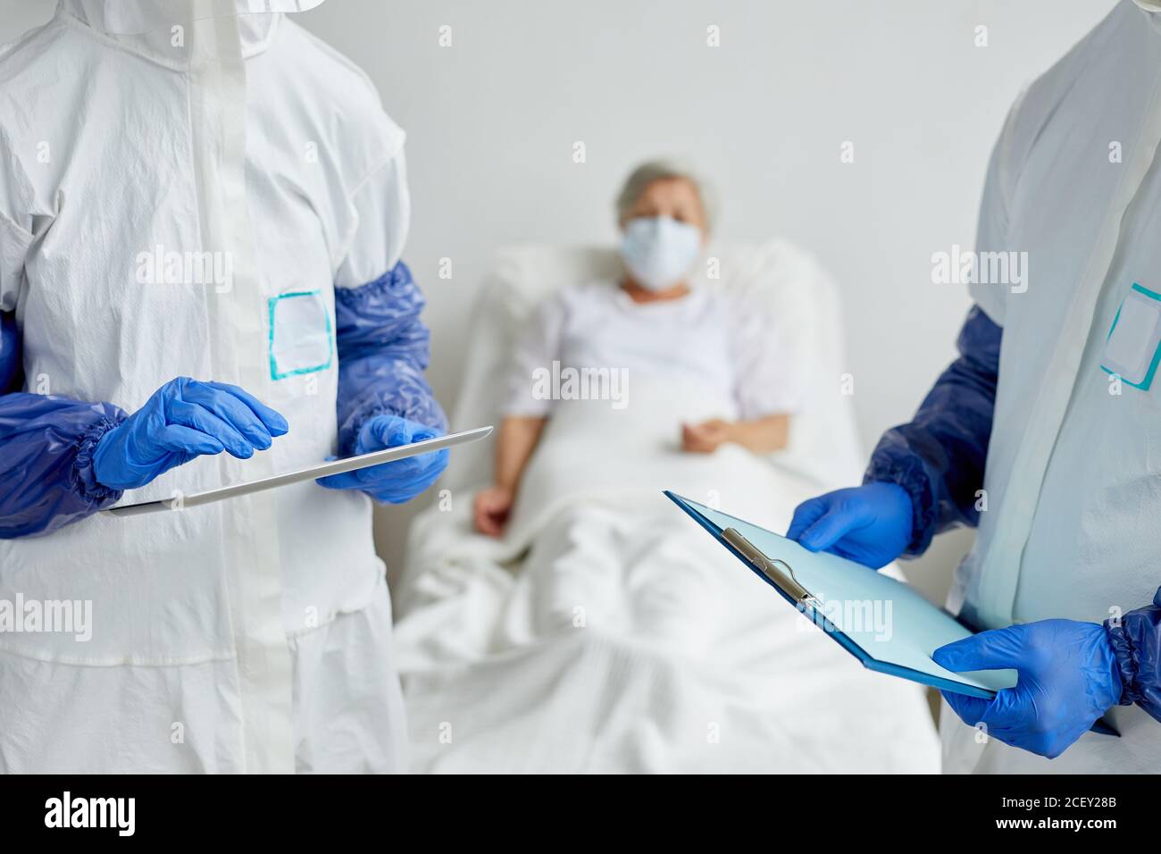 Two modern unrecognizable doctors wearing protective clothes standing in hospital ward with senior female patient on bed discussing something Stock Photo