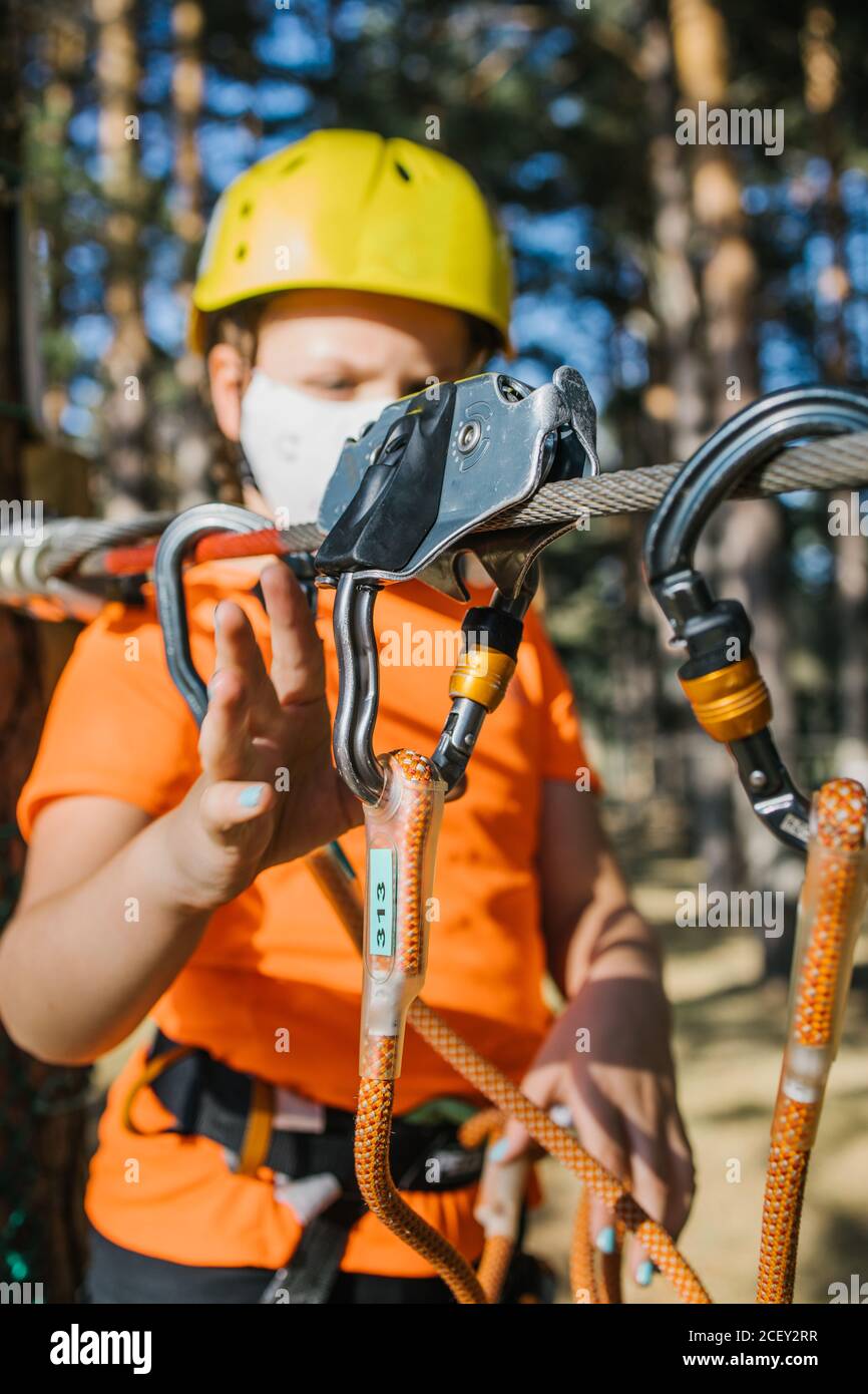 Focused girl in protective mask and helmet standing near tree in adventure park and putting carabine on safety rope during coronavirus Stock Photo
