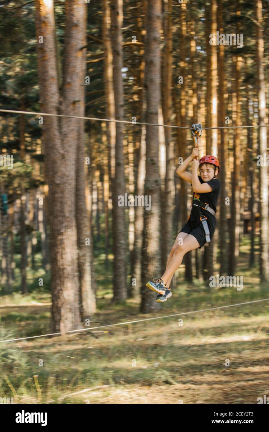 Cheerful boy in protective helmet and safety harness riding zip line and having fun in adventure park in summer Stock Photo