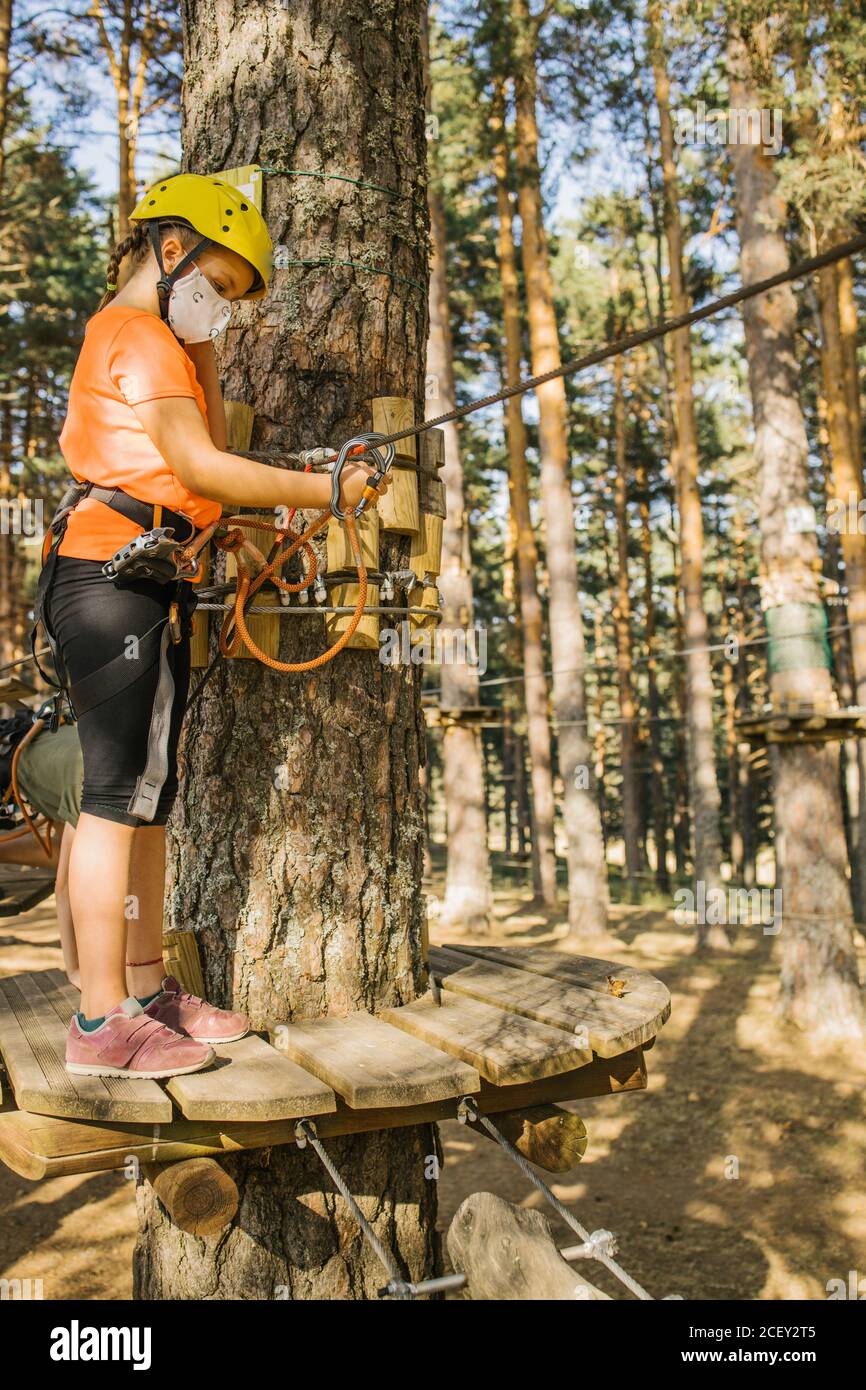 Side view of child in protective helmet and safety harness standing on wooden platform and having fun in adventure park during coronavirus Stock Photo