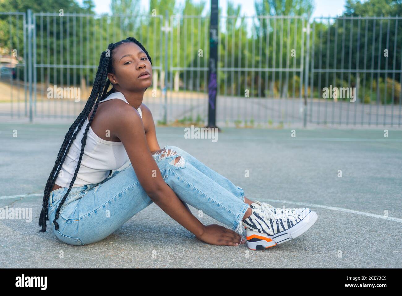 Full length of young African American female with braids wearing trendy  ripped jeans and white tank top with sneakers sitting against basketball  hoop on playground and looking at camera Stock Photo -