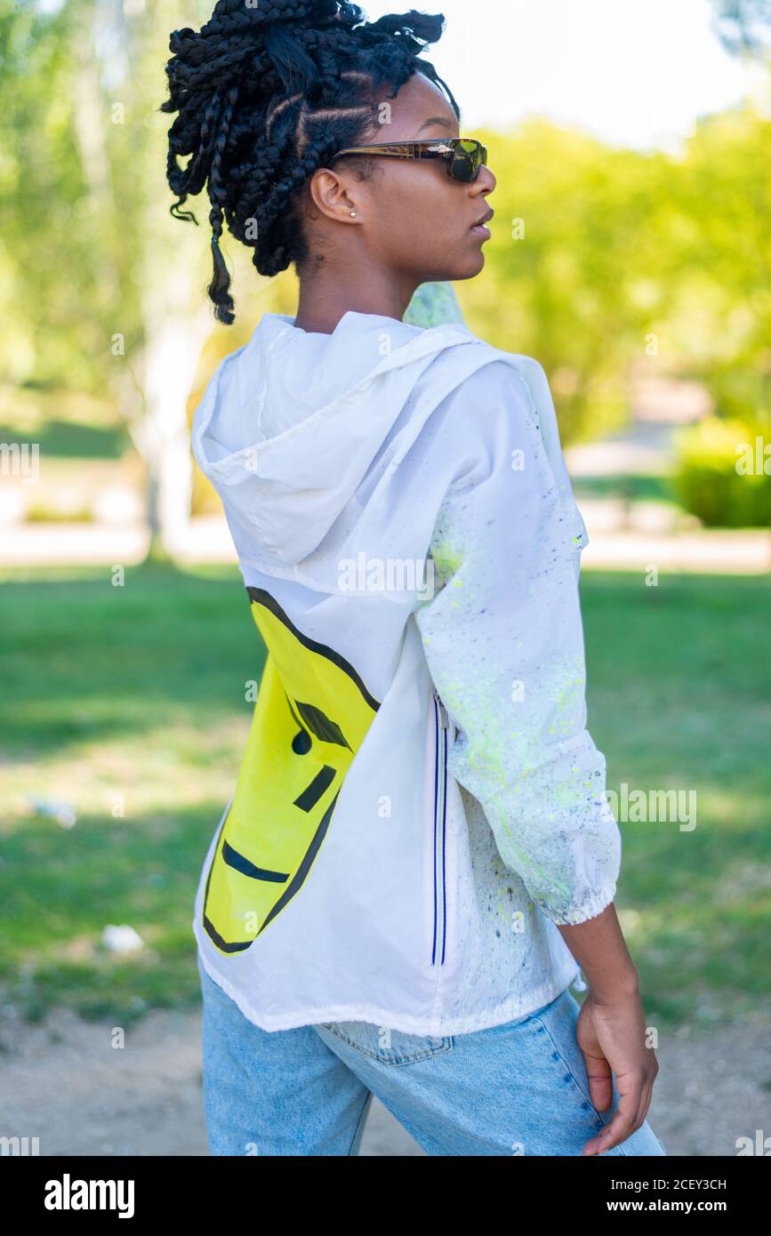 Side view of African American millennial female in sunglasses dressed in stylish white blouse over black top and trendy ripped jeans standing in green summer park Stock Photo