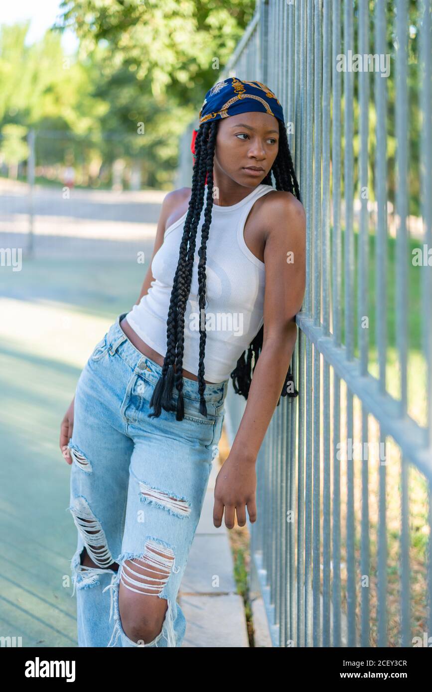 Young African American female with Afro pigtails dressed in stylish ripped jeans and white top leaning on metal fence in urban park and looking away Stock Photo