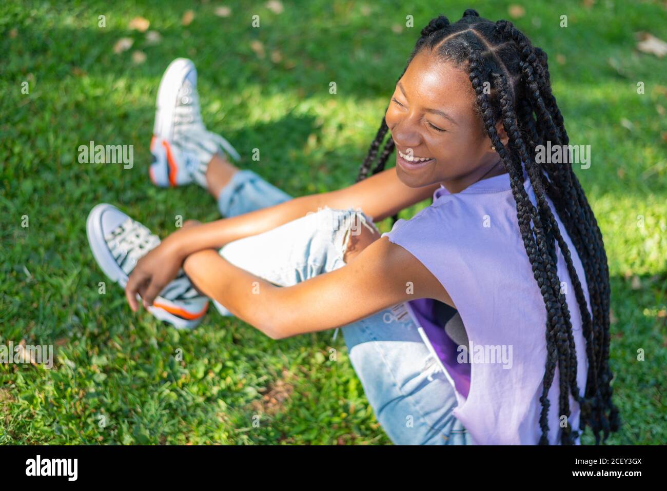 From above side view of happy young African American Woman with braids wearing casual clothes sitting on green grass and laughing while enjoying summer day in park Stock Photo